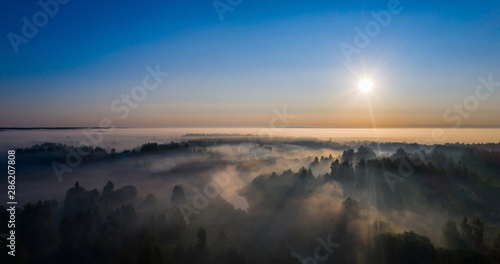 The rays of the morning sun make their way through the thick fog over the forest  field and river
