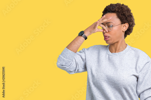 Young beautiful african american woman wearing glasses over isolated background smelling something stinky and disgusting, intolerable smell, holding breath with fingers on nose. Bad smells concept.