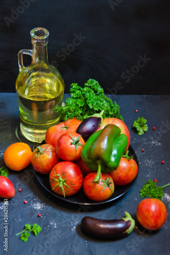 farm tomatoes of different shapes in a Cup and a bottle of olive oil sweet pepper and eggplant on a black background and parsley leaves and selective focus