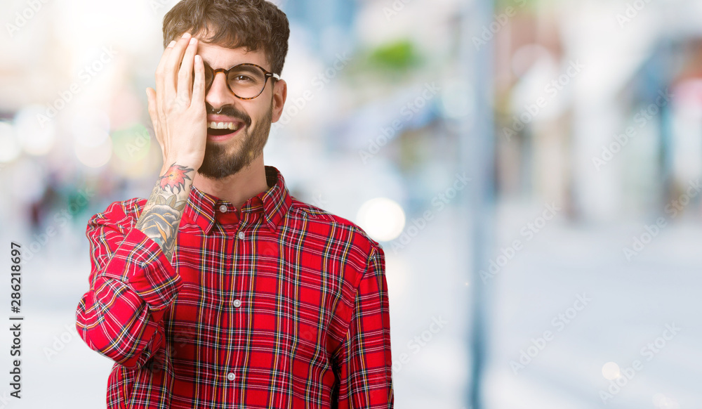 Young handsome man wearing glasses over isolated background covering one eye with hand with confident smile on face and surprise emotion. Young handsome man wearing glasses over isolated background covering one eye with hand with confident smile on face and surprise emotion.