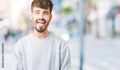 Young handsome man wearing sweatshirt over isolated background sticking tongue out happy with funny expression. Emotion concept.
