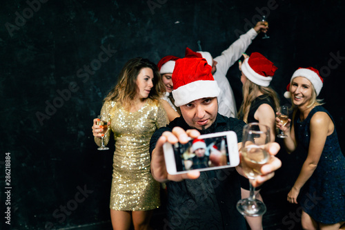 Christmas, 2020, New year party atmosphere. Happy friends in santa claus hats having fun, drinking champagne and taking selfie photos together