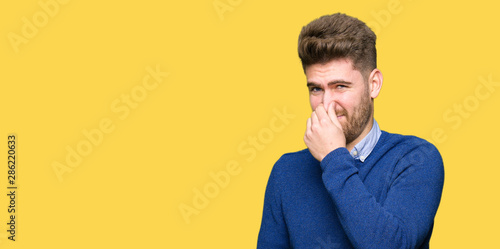 Young handsome bussines man smelling something stinky and disgusting, intolerable smell, holding breath with fingers on nose. Bad smells concept. © Krakenimages.com