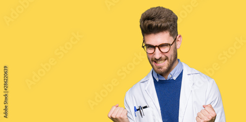 Young handsome scientist man wearing glasses very happy and excited doing winner gesture with arms raised, smiling and screaming for success. Celebration concept. photo