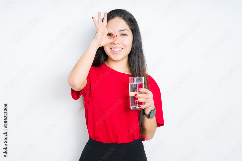 Young brunette woman drinking a glass of water over isolated background with happy face smiling doing ok sign with hand on eye looking through fingers Young brunette woman drinking a glass of water over isolated background with happy face smiling doing ok sign with hand on eye looking through fingers
