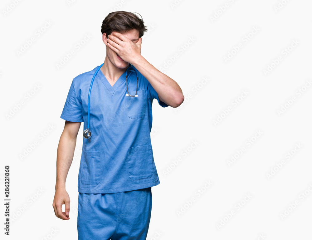 Young doctor wearing medical uniform over isolated background smiling and laughing with hand on face covering eyes for surprise. Blind concept. Young doctor wearing medical uniform over isolated background smiling and laughing with hand on face covering eyes for surprise. Blind concept.