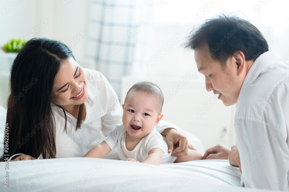 Asian father and mother are playing with new newborn baby on bed. They are smiling and warm touching to the baby with love. Parents hugging baby that crawling on the bed. Happy family concept. Asian father and mother are playing with new newborn baby on bed. They are smiling and warm touching to the baby with love. Parents hugging baby that crawling on the bed. Happy family concept.