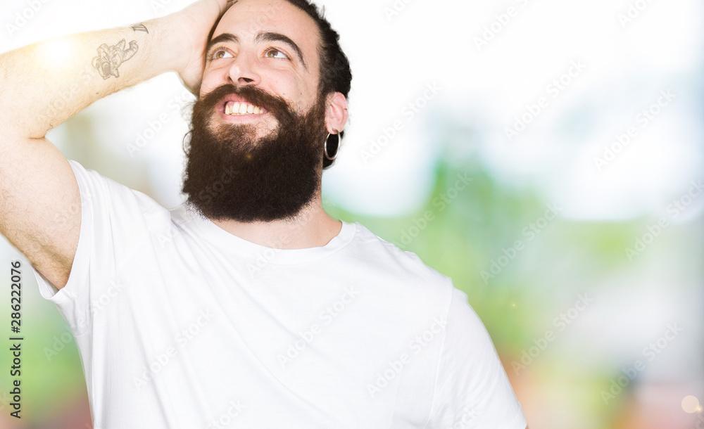 Young hipster man with long hair and beard wearing casual white t-shirt Smiling confident touching hair with hand up gesture, posing attractive Young hipster man with long hair and beard wearing casual white t-shirt Smiling confident touching hair with hand up gesture, posing attractive