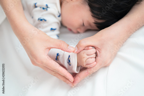 Top view of Asian mother is holding her cute sleeping newborn baby hand on bed. She making hands in hearth shape holding the hand with love in white bedroom at home. Happy family concept.