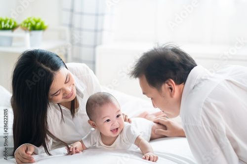 Asian father and mother are playing with new newborn baby on bed. They are smiling and warm touching to the baby with love. Parents hugging baby that crawling on the bed. Happy family concept.