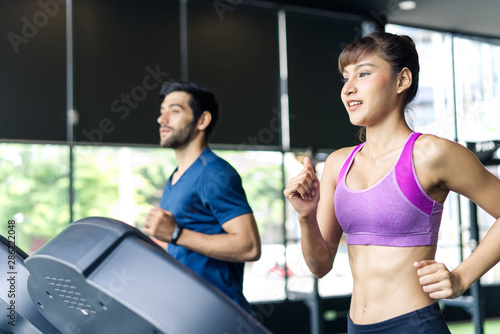 Fototapeta Naklejka Na Ścianę i Meble -  Young fit beautiful Asian woman and handsome Caucasian man running on treadmill or running machine in modern fitness gym. Seen from side view while they focusing on running. Workout in Gym and fitness