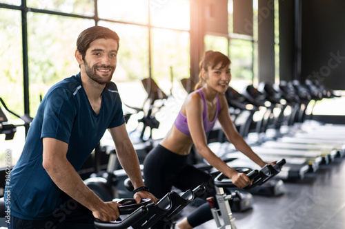 Young fit handsome Caucasian man and beautiful Asian woman cycling on bike machine in modern fitness gym. Seen from side view while they focusing on exercising. Workout in Gym and fitness for health.