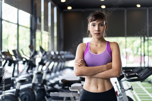 Portrait of young pretty Asian girl with six packs in purple color sportswear standing and crossing arms in gym or fitness club. Behind her is exercise machines and equipment. Fitness & Gym concept.