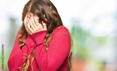 Young beautiful woman wearing red sweater with sad expression covering face with hands while crying. Depression concept.