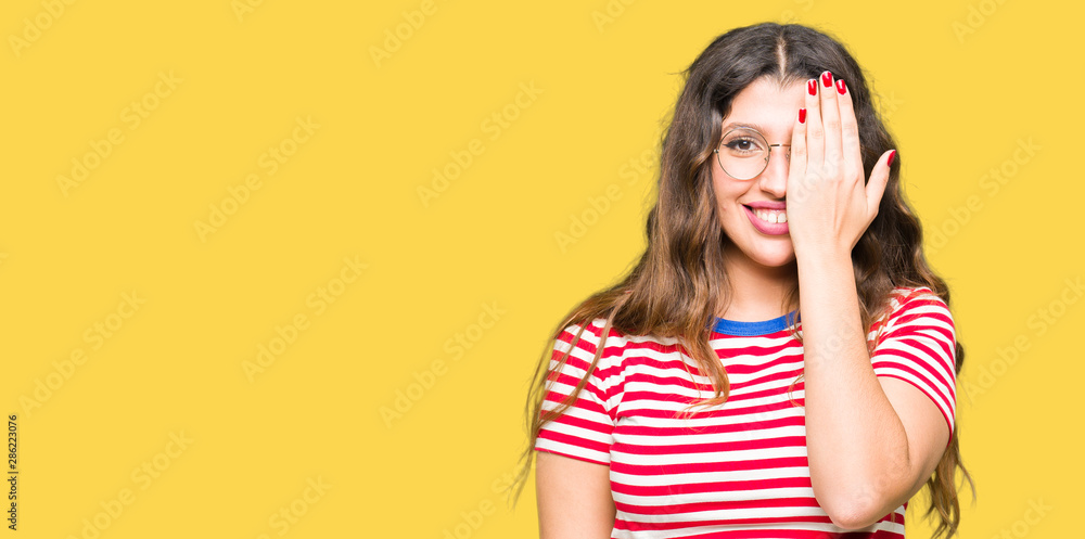 Young beautiful woman wearing glasses covering one eye with hand with confident smile on face and surprise emotion. Young beautiful woman wearing glasses covering one eye with hand with confident smile on face and surprise emotion.