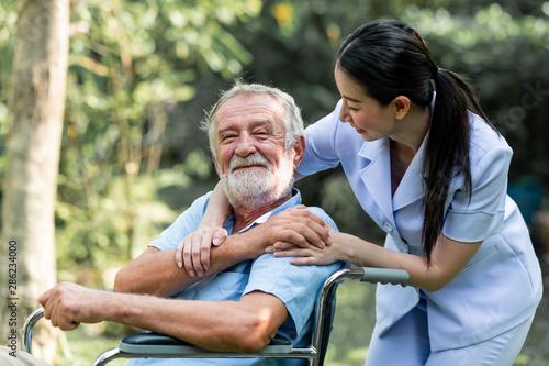 Caring nurse with senior man sitting on wheelchair in gaden. Asian woman, caucasian man. Happy feeling.