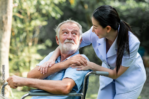 Caring nurse with senior man sitting on wheelchair in gaden. Asian woman, caucasian man. Looking at nurse.