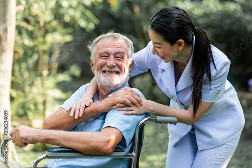 Caring nurse with senior man sitting on wheelchair in gaden. Asian woman, caucasian man. Big smile.