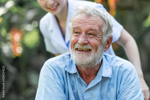 Caring nurse helping senior man sitting on wheelchair in gaden. Asian woman, caucasian man. Happy smile.