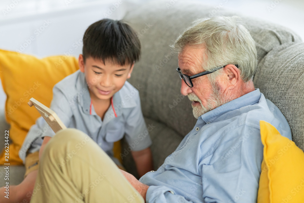 Fototapeta premium Grandfather with child reading. Senior caucasian male with asian boy. Reading book together in living room at home. Educating him.