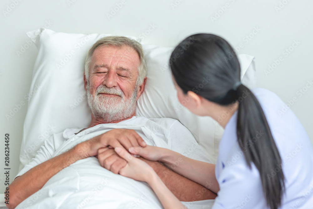 Fototapeta premium Senior man in bed with nurse in retirement home. Caucasian male with asian woman. Reassuring pose.