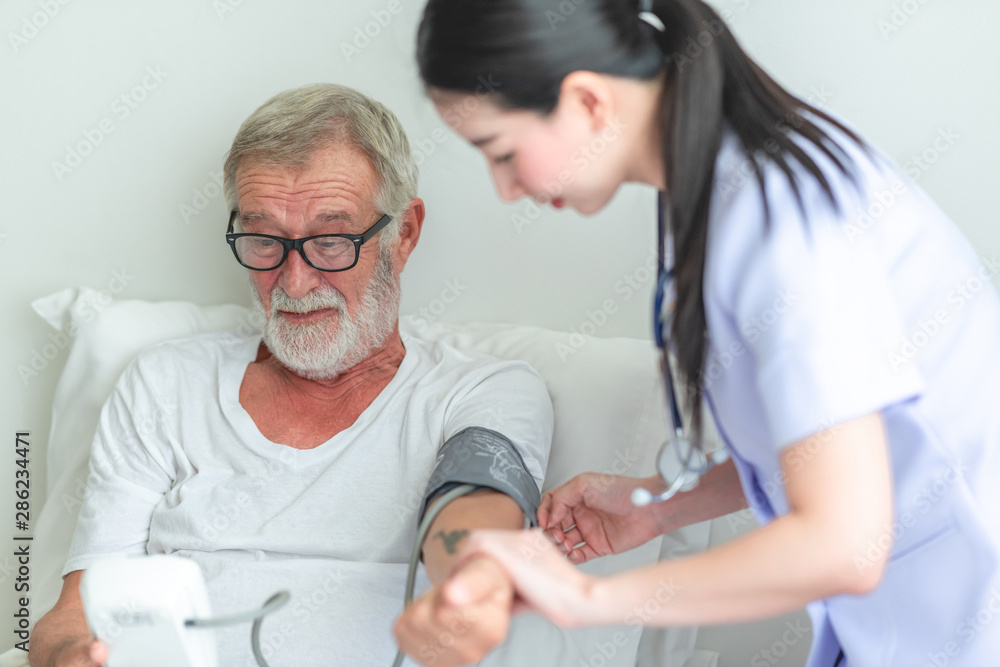 Obraz premium Senior man in bed with nurse in retirement home. Caucasian male with asian woman. Checking result of blood pressure.