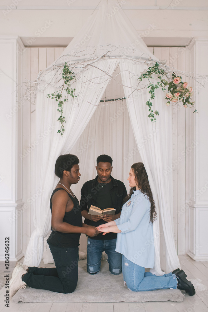 Fotka „Interracial couple standing on knees in front of wedding arch. Interracial bridal ceremony. Caucasian woman getting married with her african dark skinned boyfriend. Indoor amateur wedding ceremony.“ ze služby Stock |