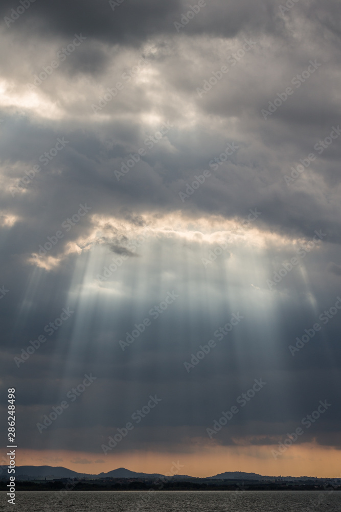 Sunrays at near sunset, with dark clouds in the background, an orange sky, and Trasimeno lake (Umbria, Italy) below