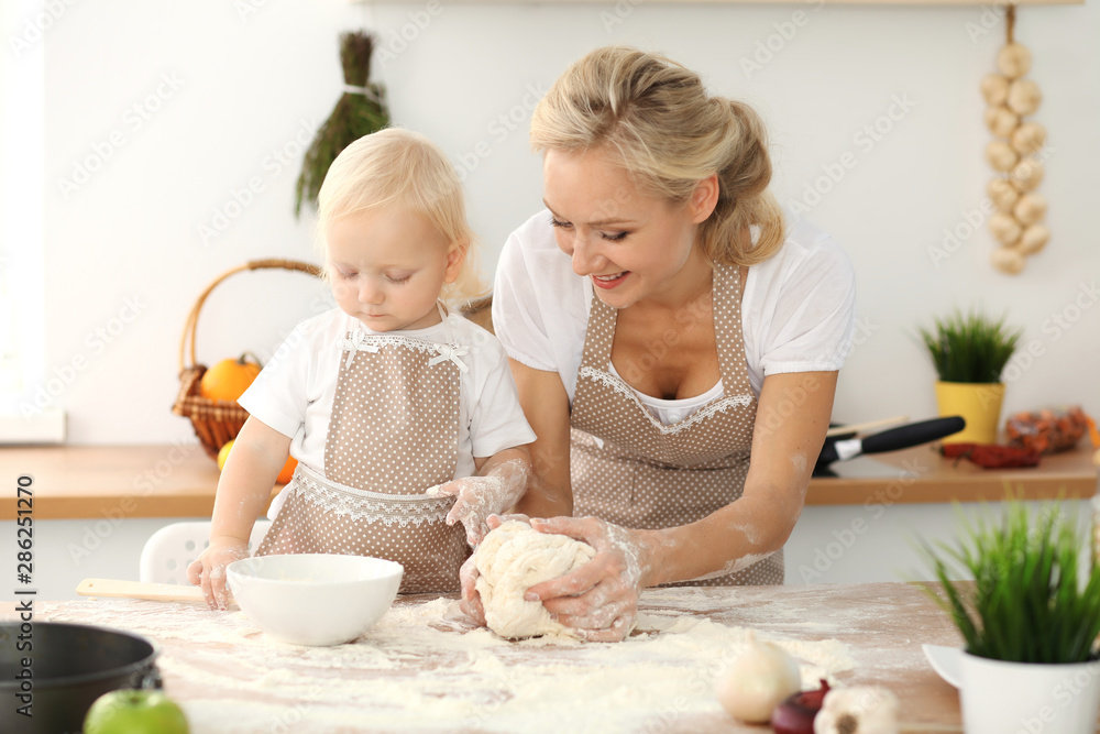 Little girl and her blonde mom in beige aprons playing and laughing while kneading the dough in kitchen. Homemade pastry for bread, pizza or bake cookies. Family fun and cooking concept Little girl and her blonde mom in beige aprons playing and laughing while kneading the dough in kitchen. Homemade pastry for bread, pizza or bake cookies. Family fun and cooking concept