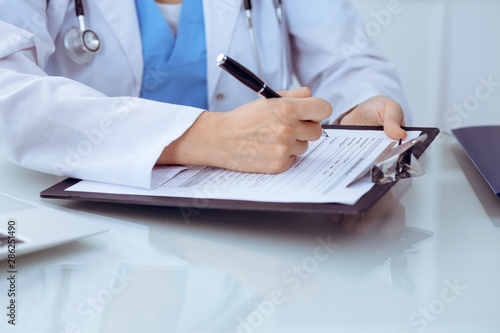 Doctor woman filling up medical form while sitting at the table, close-up of hands