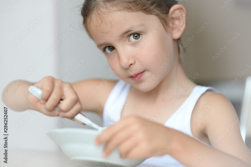 beautiful little caucasian girl in white tank top eating breakfast holding plate and spoon sitting in bright kitchen beautiful little caucasian girl in white tank top eating breakfast holding plate and spoon sitting in bright kitchen