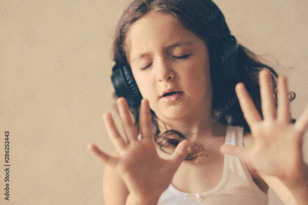 delightful little curly girl with closed eyes wearing headphones listening sound of music with hand gesture
