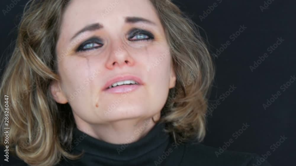Beautiful young tearful unhappy woman crying, washing off makeup. Real tears running down on face that screams of desperation in hysterics in a black jacket on a black background, face close up view