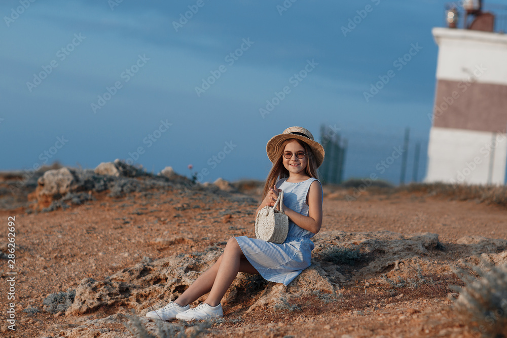 A girl of 10-11 years spends time alone on a rocky shore near the blue ocean on open air in the summer. Cute girl in a straw hat on a rocky coast. Little girl in a blue dress on a background of blue s