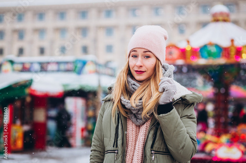 A girl in a pink hat and a knitted woolen scarf walks at the Christmas market. blonde girl smiles and laughs on the background of the winter fair. © Stanislav