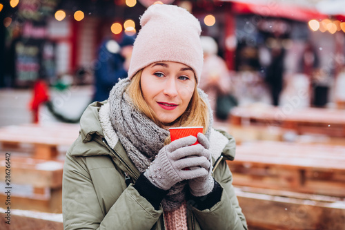 Girl in a pink hat and a knitted woolen scarf drinks cocoa at the Christmas market. woman with a red glass on a city holiday. girl basks in hot tea on the background of the Christmas market. © Stanislav