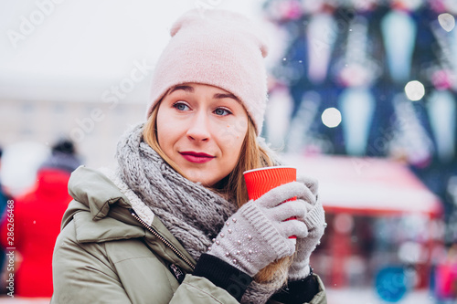 Girl in a pink hat and a knitted woolen scarf drinks cocoa at the Christmas market. woman with a red glass on a city holiday. girl basks in hot tea on the background of the Christmas market. © Stanislav