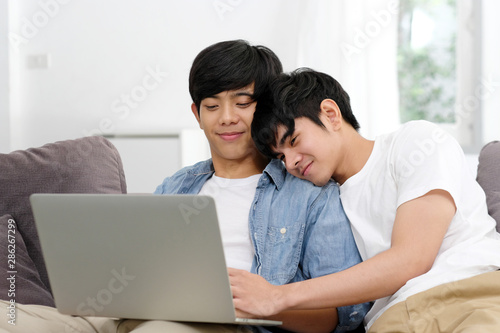 Happy young asian gay man couple using laptop computer while sitting onsofa at home, homosexual and lgbt with technology lifestyle concept © mangpor2004