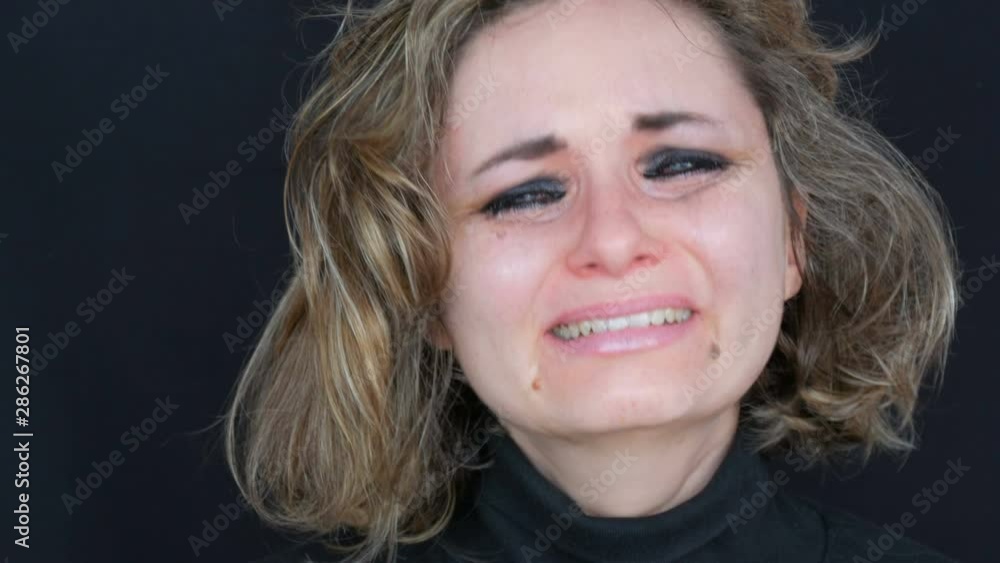 Beautiful young tearful unhappy woman crying, washing off makeup. Real tears running down on face that screams of desperation in hysterics in a black jacket on a black background, face close up view Beautiful young tearful unhappy woman crying, washing off makeup. Real tears running down on face that screams of desperation in hysterics in a black jacket on a black background, face close up view