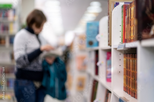 Photo of shelves of book in bookshop or store, library, natural blurred by optic lense with reading woman on background.