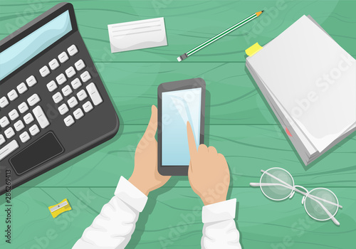 Moody flat lay background with workplace. Light green wooden background with stack of paper, eye glasses, pencil and laptop. A man holding a smartphone.