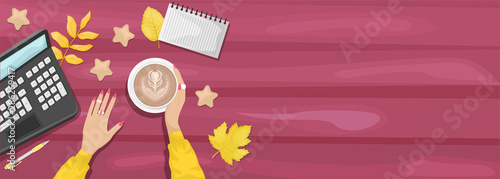 Autumn flat lay background with workplace in a cafe. Women's hands holding a Cup of cappuccino. Burgundy wooden background with yellow autumn leaves. Laptop, notebook, pen. Place for text.