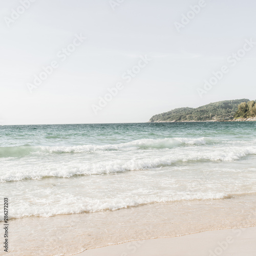 Beautiful tropical beach view with white sand, blue sea with waves and green island on the horizon on Phuket, Thailand. Minimal composition with neutral colors. Summer concept. Natural background.