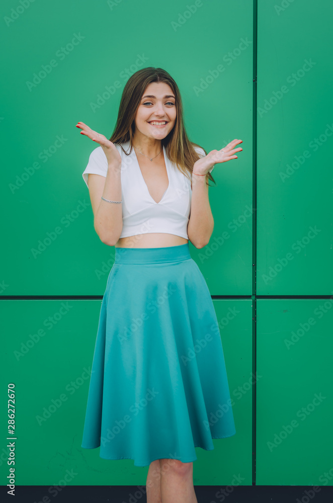 Surprised women dressed in white topic and turqoise skirt looking at camera near turqoise wall Surprised women dressed in white topic and turqoise skirt looking at camera near turqoise wall