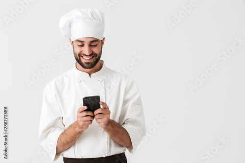 Cheerful smiling young chef posing isolated over white wall background in uniform using mobile phone.