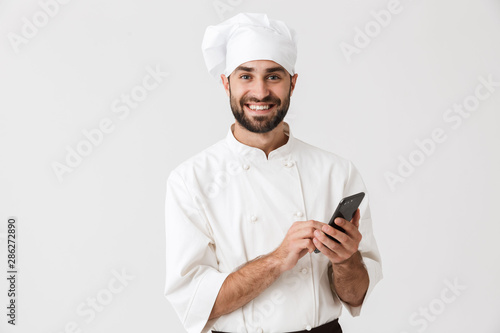 Cheerful smiling young chef posing isolated over white wall background in uniform using mobile phone. © Drobot Dean