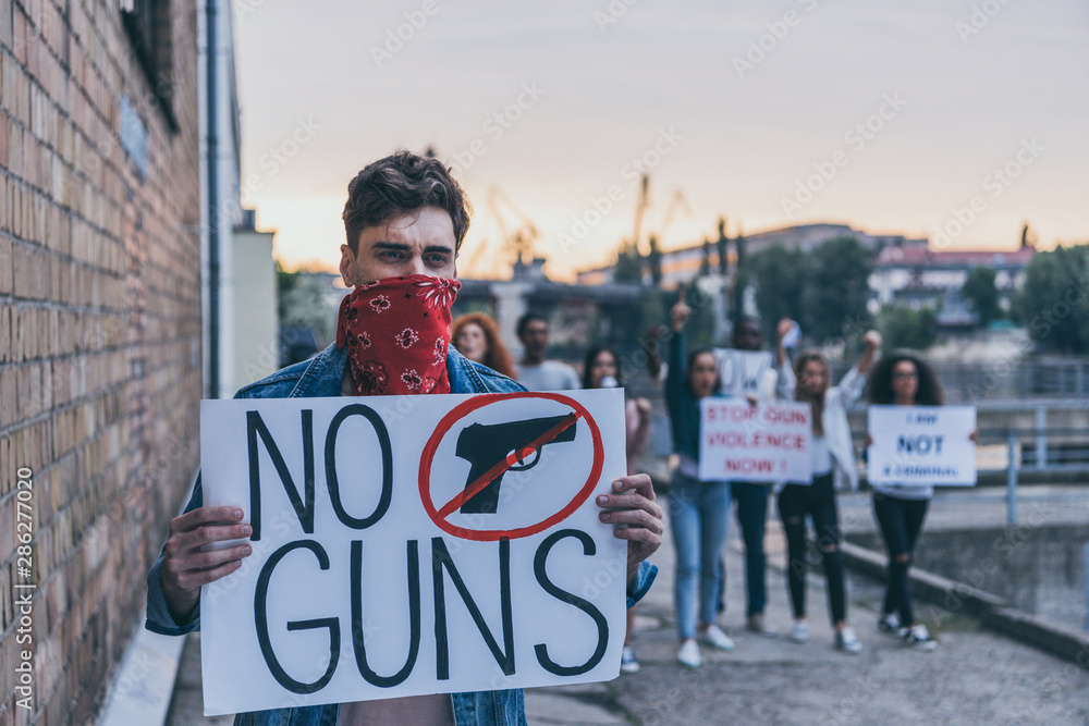 selective focus of man with scarf on face holding placard with no guns lettering near multicultural people selective focus of man with scarf on face holding placard with no guns lettering near multicultural people