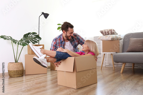 Young married couple moving into a new home. Attractive blonde woman sitting in cardboard box, bearded man pushes her. Newely weds fooling around. Minimal interior background, copy space, close up photo