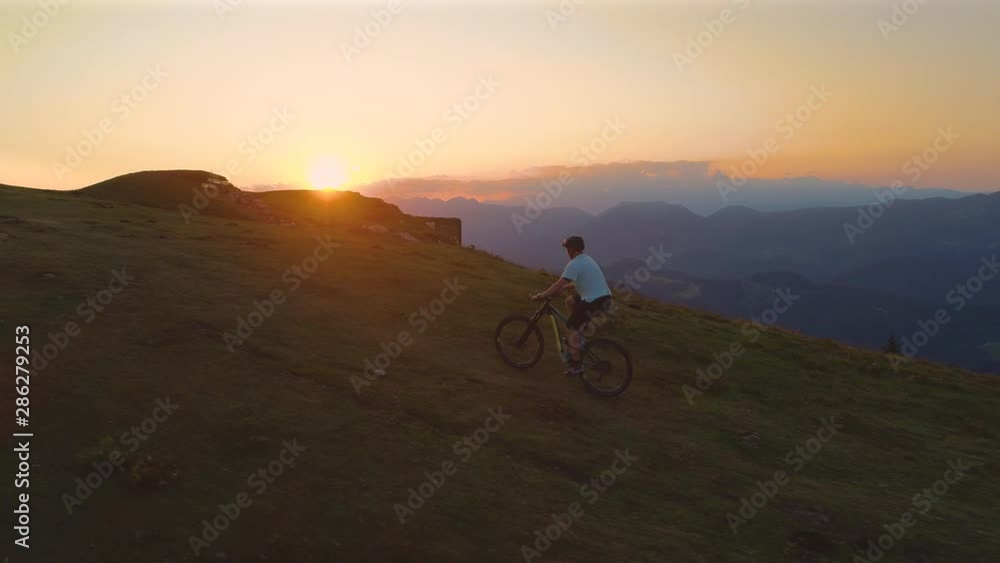 AERIAL: Unrecognizable mountain biker riding his bicycle up a grassy hill at picturesque sunset. Young male tourist enjoying a scenic mountain bike ride on an ebike on a beautiful summer morning. AERIAL: Unrecognizable mountain biker riding his bicycle up a grassy hill at picturesque sunset. Young male tourist enjoying a scenic mountain bike ride on an ebike on a beautiful summer morning.