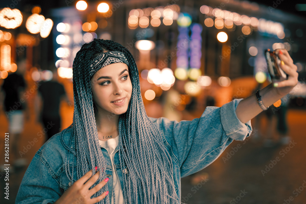 Young unconventional woman with blue pigtails taking selfie by smartphone in night city